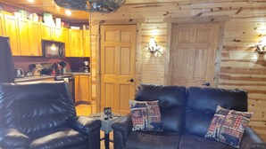 A cozy living room with a leather sofa and armchair, wooden walls, and a kitchen area with wooden cabinets and a microwave. - River Ridge Cottage (Spooner)