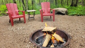 A campsite with two red Adirondack chairs, a small wooden table, and a metal fire pit with burning wood. - River Ridge Cottage (Spooner)