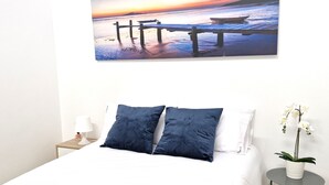 Een netjes opgemaakt bed met witte lakens en twee donkerblauwe kussens, een witte handdoek en een lijst met een afbeelding van een strandscène aan de muur boven het bed.