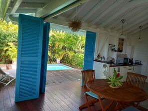 A wooden deck with a pool, blue shutters, a dining table with chairs, and a kitchen area with a hanging light fixture. - Courbaril villas, Two bungalows with private pools and sea view (st louis)
