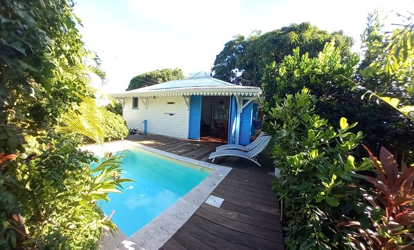 A pool area with a wooden deck, a small white building with blue shutters, and lush greenery surrounding the space.