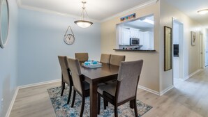 A dining area with a wooden table, four chairs, a hanging light fixture, a wall clock, and a kitchen visible through an archway. - LOCATION**LOCATION**LOCATION (Gulf Shores)