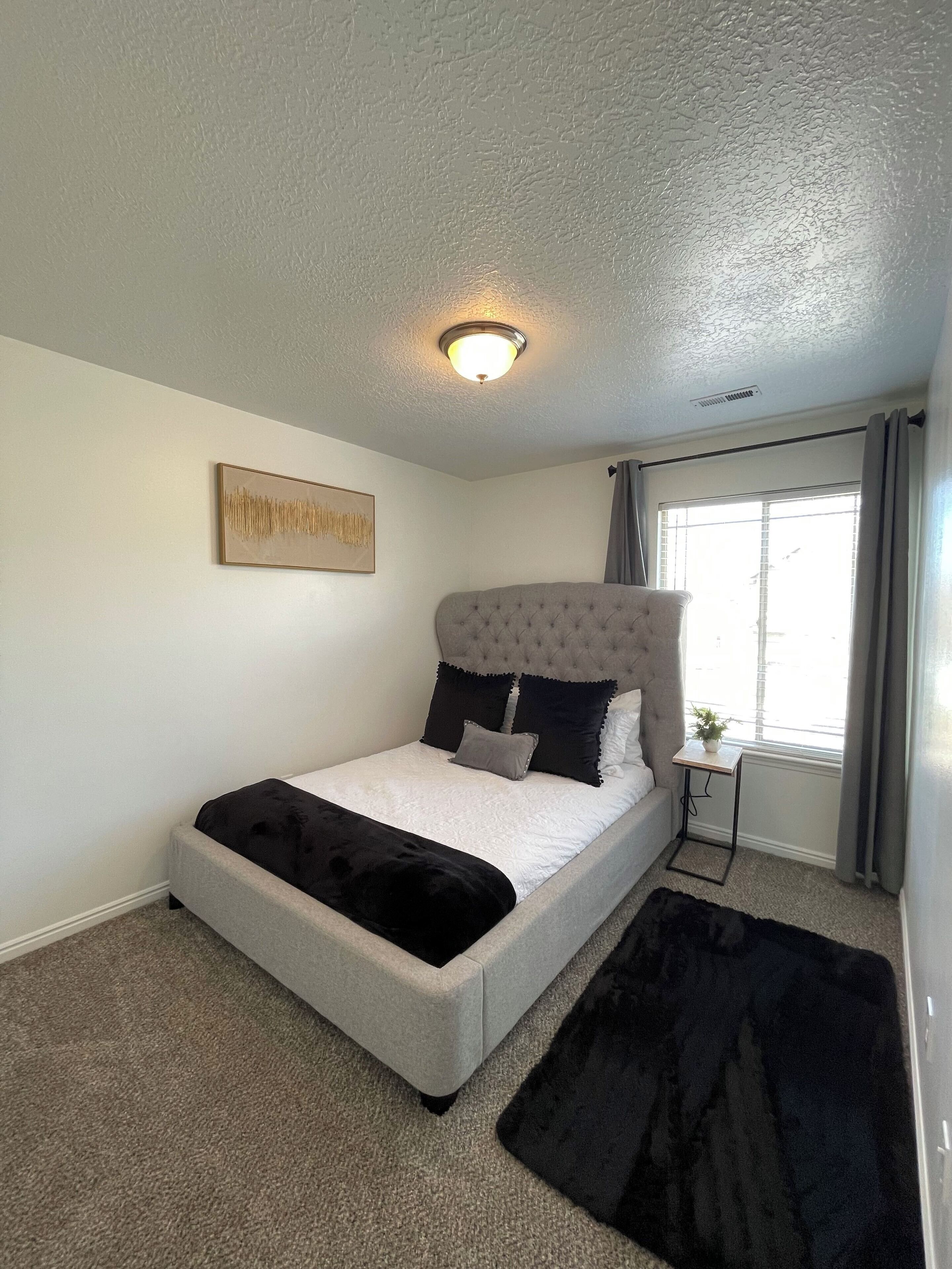 Une chambre à coucher avec un lit, une petite table de chevet, une fenêtre avec des rideaux et un luminaire au plafond.