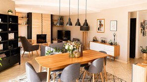 A modern living room with a wooden dining table, grey chairs, a black bookshelf, a flat-screen TV, and pendant lights.