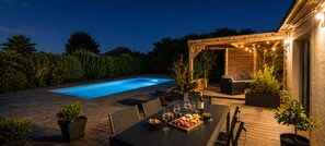A poolside area with a wooden deck, outdoor seating, and a pergola with string lights.