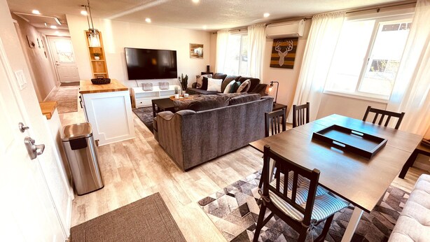 A modern living room with a grey sofa, a dining table with chairs, a TV, and a kitchen area with a fridge and a sink. - Westside ^Cottage^ (Redding)