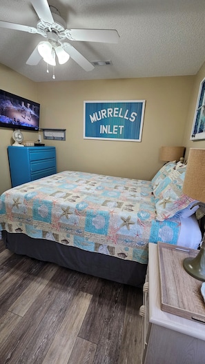 A bedroom with a bed, a ceiling fan, a TV, a blue dresser, and a sign that reads "MURRELLS INLET".