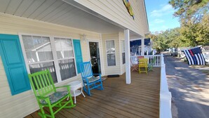 A covered porch with wooden flooring, a white railing, and colorful Adirondack chairs.
