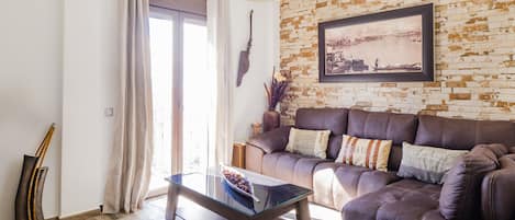 A living room with a brown leather sofa, a glass coffee table, and a brick-patterned wall with a framed picture.