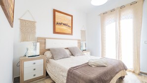 A bedroom with a bed, bedside tables, a nightstand, a wall-mounted lamp, a macrame wall hanging, and a painting of a desert scene.