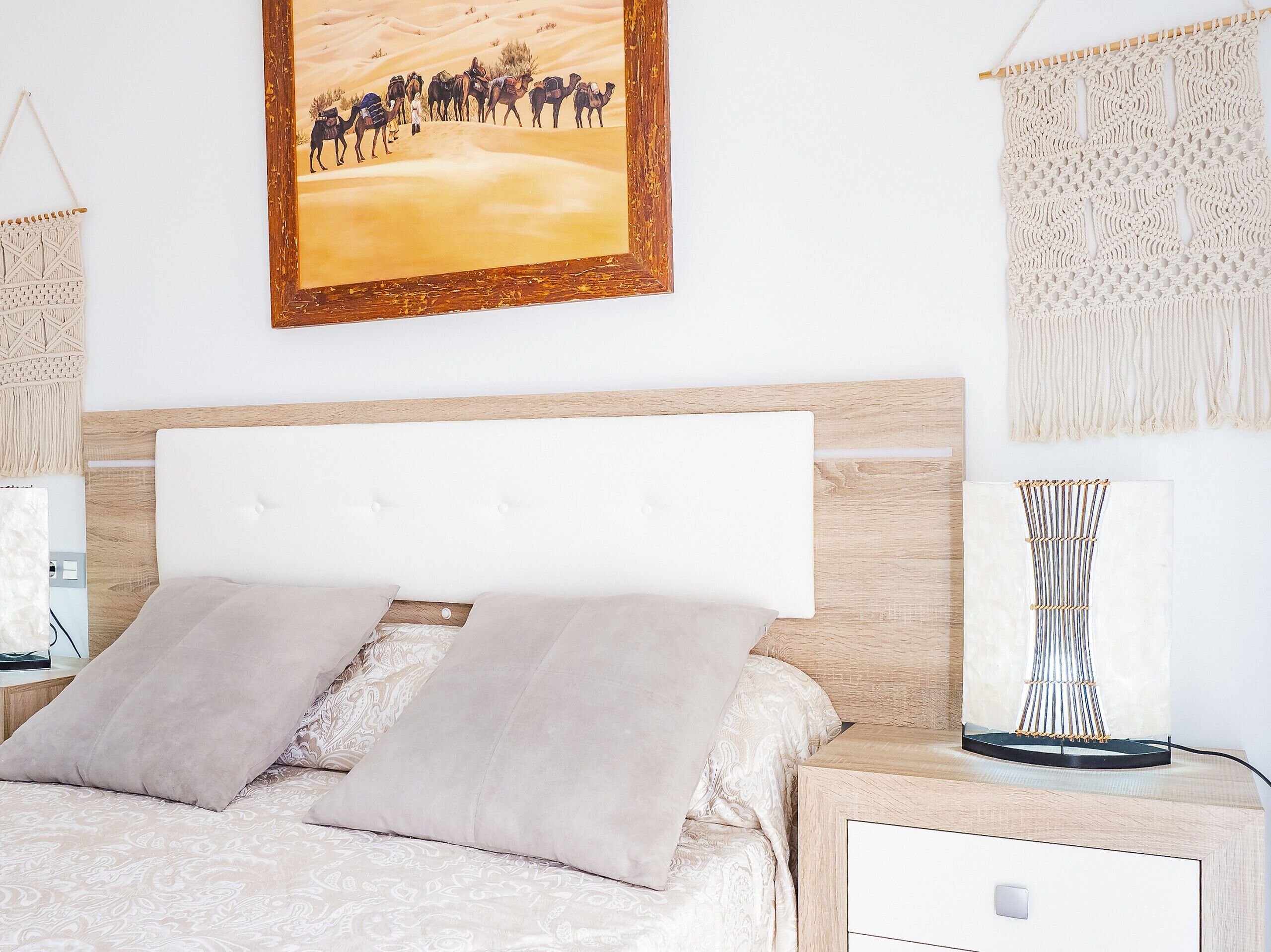 Un dormitorio con cama, mesita de noche, lámpara, un tapiz de macramé y un cuadro con camellos.