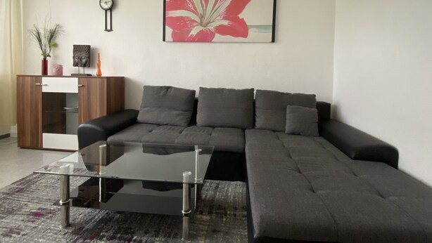 Una sala de estar moderna con un sofá modular, una mesa de centro de vidrio, un reloj y un cuadro grande con motivos florales en la pared.