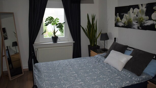 A bedroom with a bed, a window with curtains, a potted plant, a radiator, a mirror, and a painting on the wall. - Vacation home Zum Unneschmitz, near the Rhine/Moselle, (Udenhausen)