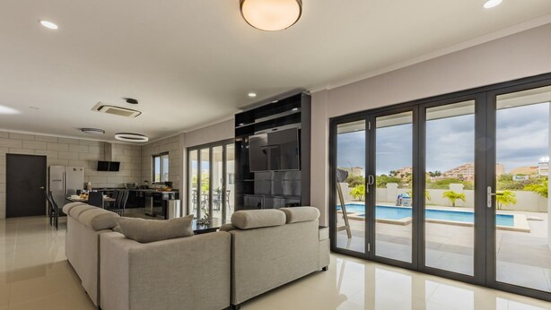 A modern living room with a sofa, a dining area, and a kitchen. There is a large sliding glass door leading to a pool area. - NEW 6BR Villa w/ Pool across street @ PalmBeach (Noord)