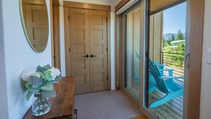 A hallway with a wooden door, a glass door leading to an outdoor area with a blue chair and table, and a vase with flowers on a wooden table.