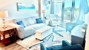 Un salon comprenant un canapé, un pouf et des fauteuils, une table basse, une lampe et un tableau représentant une scène de plage.