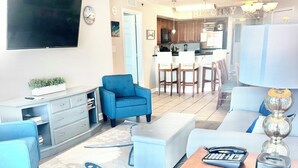 Ett modernt vardagsrum med en platt-TV, en blå fåtölj, ett soffbord och en matplats med trästolar och en kristallkrona.