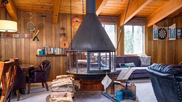 Interior acolhedor de cabana com lareira a lenha, estantes de livros, móveis de couro e um alvo de dardos.