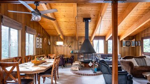 Interior em madeira com sala de jantar, lareira e sala de estar com sofá e poltronas.