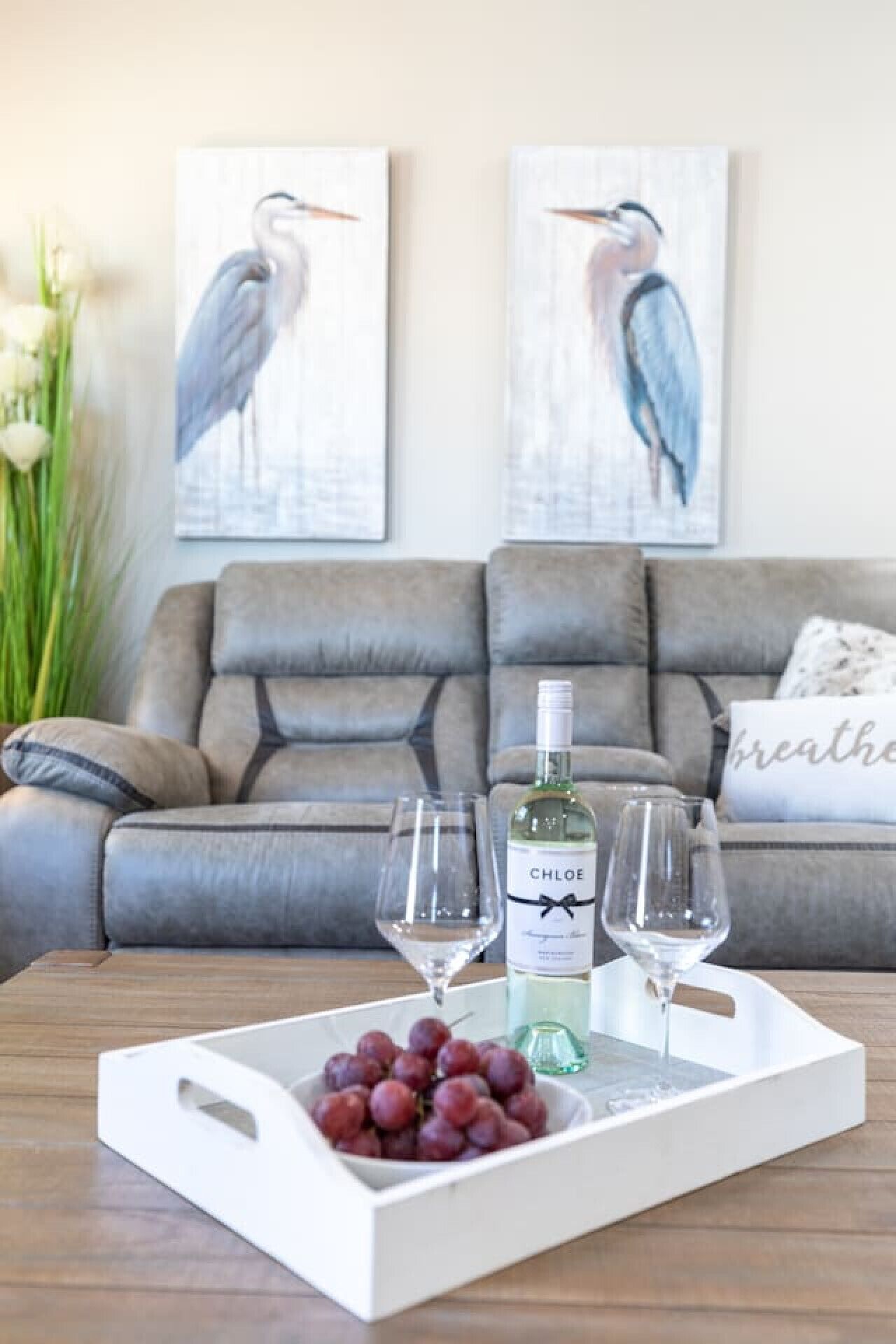 A living room with a grey sofa, two framed bird paintings, a white tray with a bottle of Chloe wine, two glasses, and a bowl of grapes on a wooden coffee table.