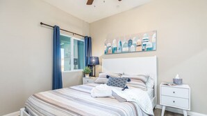 Um quarto com cama, mesa de cabeceira, luminária, janela com cortinas e um quadro na parede.