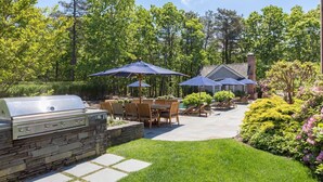 An outdoor patio with a grill, dining area, and umbrellas. - Chic Hamptons Retreat in Watermill with Tennis, Pool, Spa, Steam Room, and Gym (Water Mill)