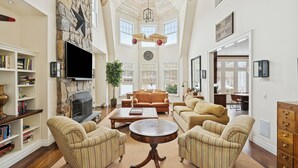 A spacious living room with a stone fireplace, a large flat-screen TV, and a cozy seating area with striped sofas and a coffee table. - Chic Hamptons Retreat in Watermill with Tennis, Pool, Spa, Steam Room, and Gym (Water Mill)