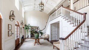 A spacious foyer with a grand staircase, a round table with chairs, and a large potted plant. - Chic Hamptons Retreat in Watermill with Tennis, Pool, Spa, Steam Room, and Gym (Water Mill)