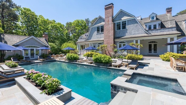 A pool area with lounge chairs and umbrellas, surrounded by a large house with a brick chimney. - Chic Hamptons Retreat in Watermill with Tennis, Pool, Spa, Steam Room, and Gym (Water Mill)