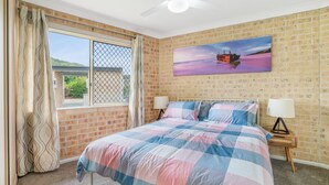 A bedroom with a brick wall, a large window with curtains, a bed with a checkered bedspread, two bedside lamps, and a painting of a ship on the wall.