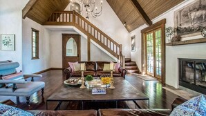 A spacious living room with a high ceiling, a fireplace, a staircase, and a large window.