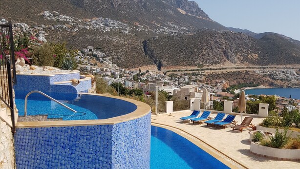 Una piscina con borde de azulejos, sillas de descanso y vista a un pueblo costero y montañas al fondo.