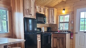 Una cocina tipo cabaña con armarios de madera, un refrigerador negro, un microondas y una estufa. Hay una ventana con persianas, un lavamanos y una puerta.