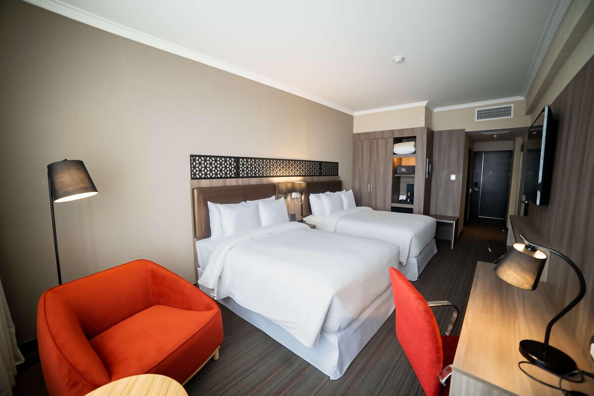 A hotel room with two beds, a desk, a chair, a lamp, and a TV.
