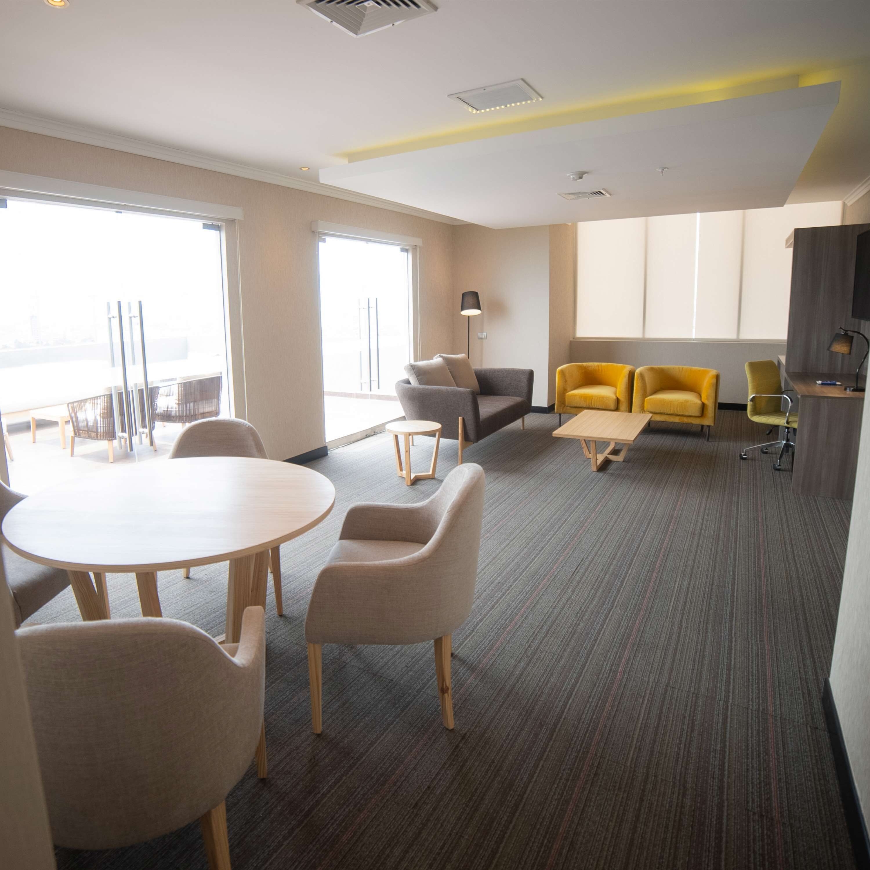 A modern hotel room with a large window, a round table with chairs, a sofa, and a small desk with a lamp.