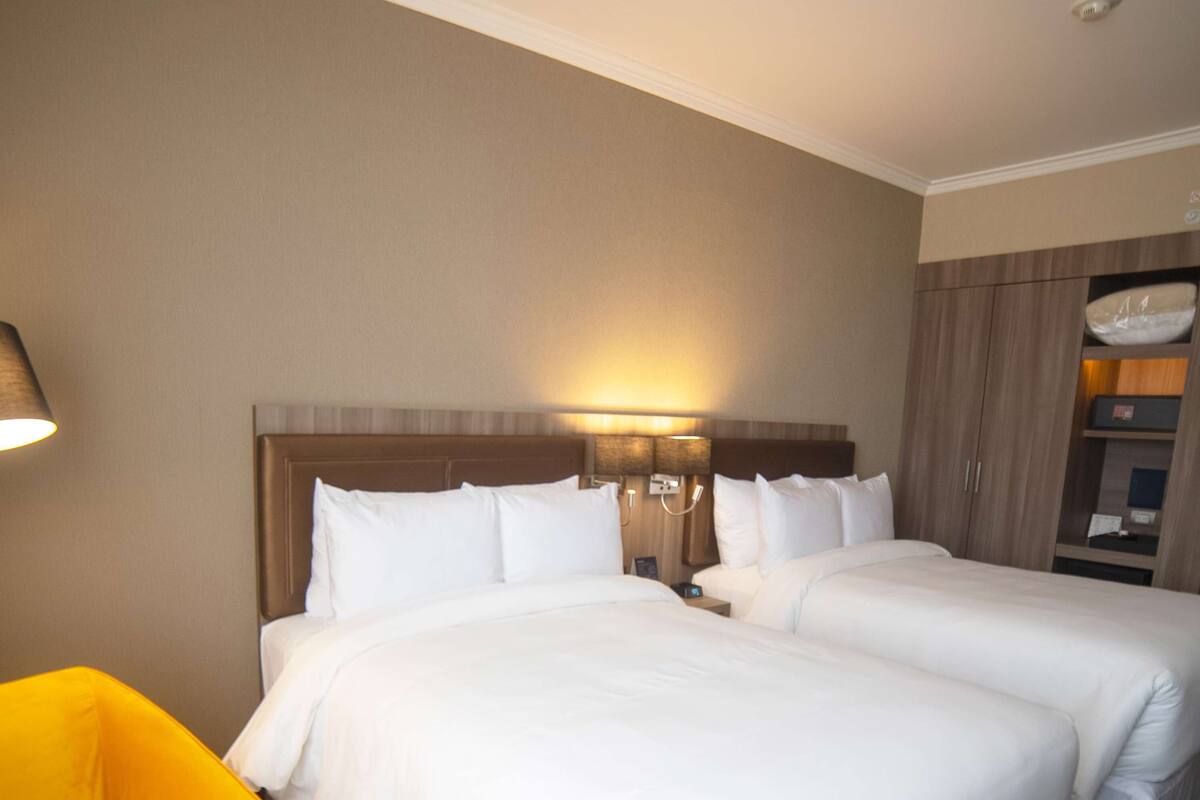 A hotel room with two beds, a nightstand, a lamp, and a yellow chair.