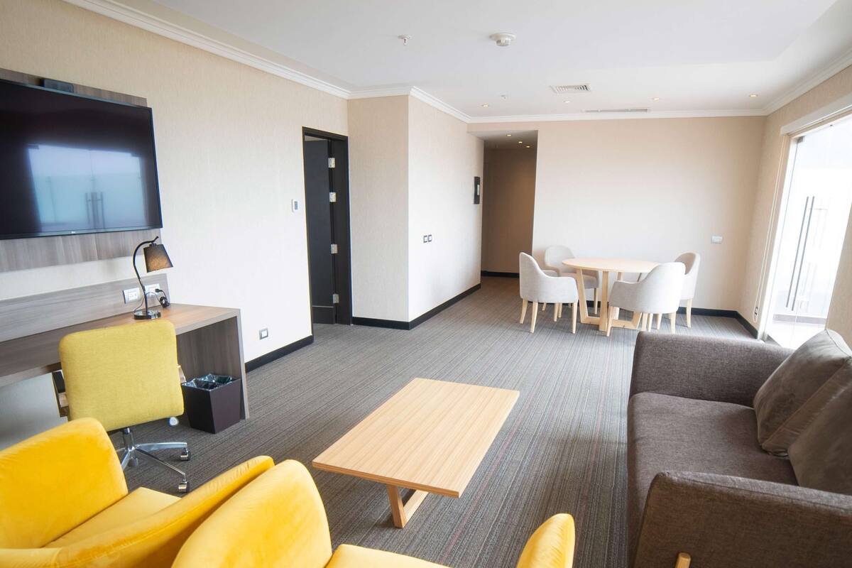A modern hotel room with a flat-screen TV, a wooden coffee table, yellow and brown seating, a small dining table with chairs, and a door leading to another room.