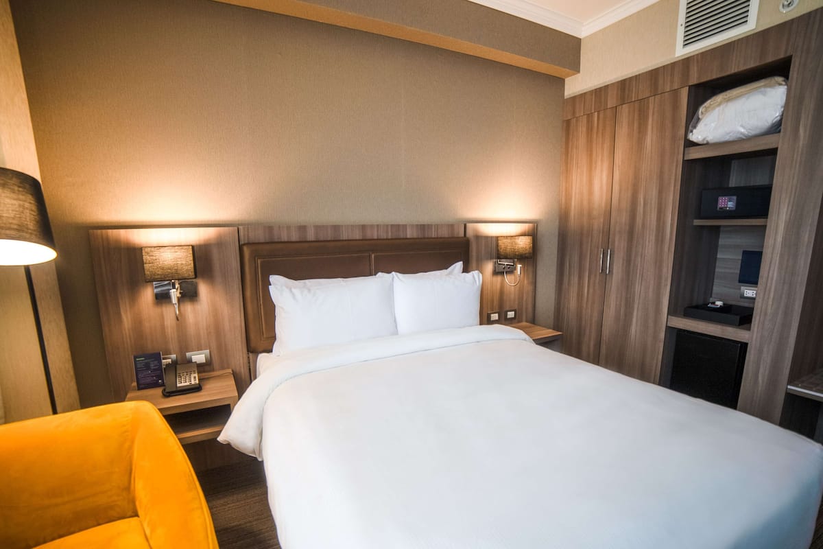 A hotel room with a large bed, two bedside tables with lamps, a wooden wardrobe, and a yellow armchair.