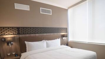 Una habitación de hotel con una cama grande, cabecero de madera con panel decorativo, dos lámparas de noche, mesita de noche y una ventana con persianas.
