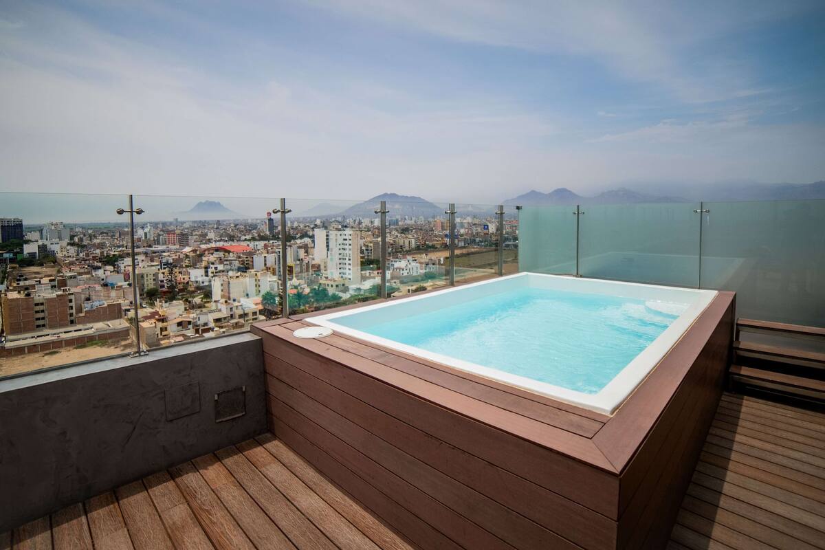A rooftop pool with a wooden deck, surrounded by city buildings and mountains in the background.