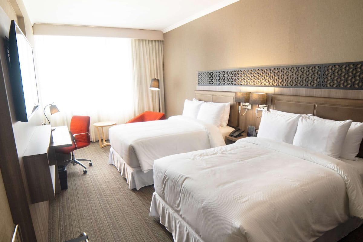 A hotel room with two beds, a desk, a chair, a lamp, and a view of the outdoors.