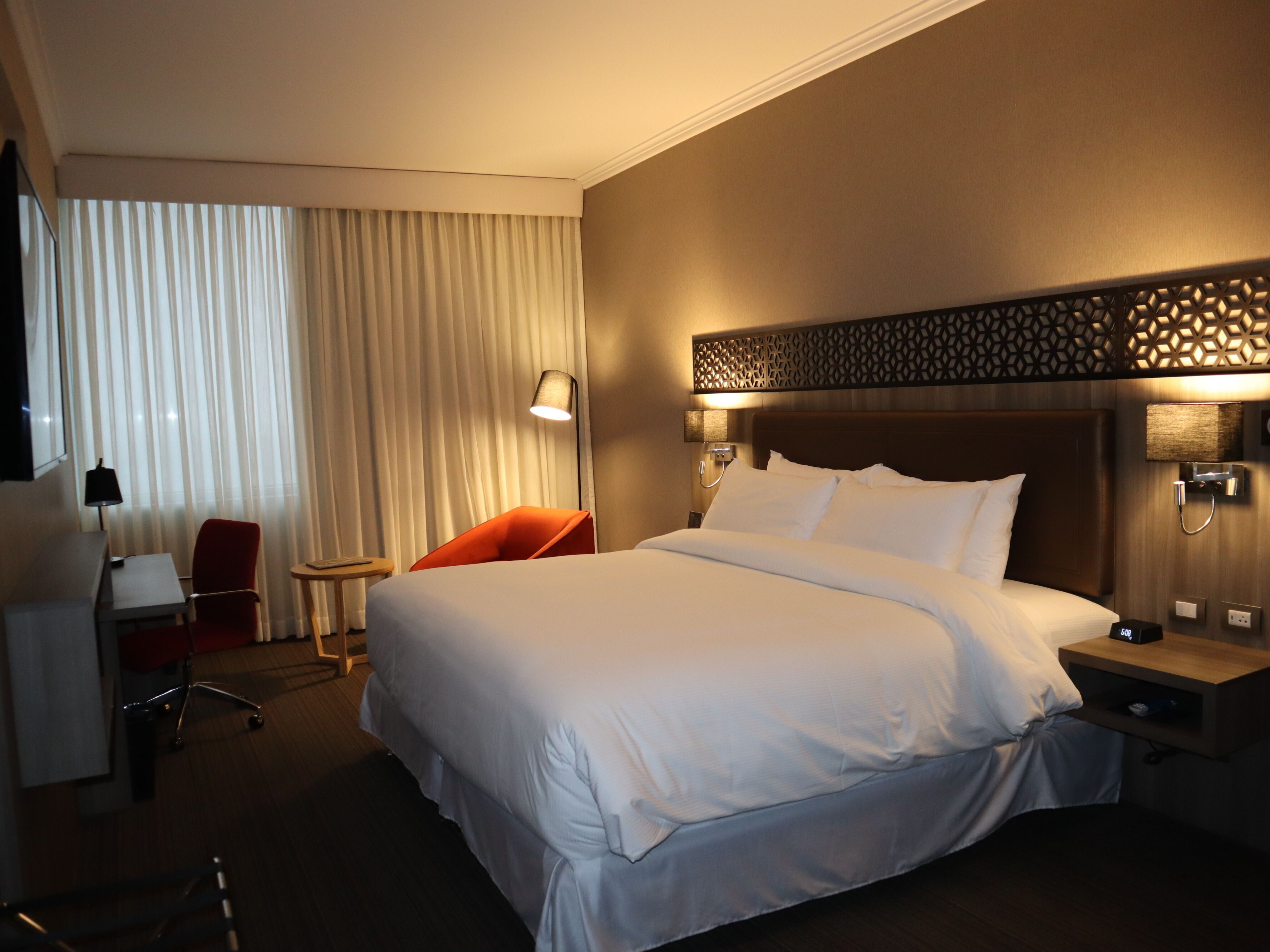 Una habitación de hotel con una cama grande, un escritorio, una silla y un ventanal con cortinas.