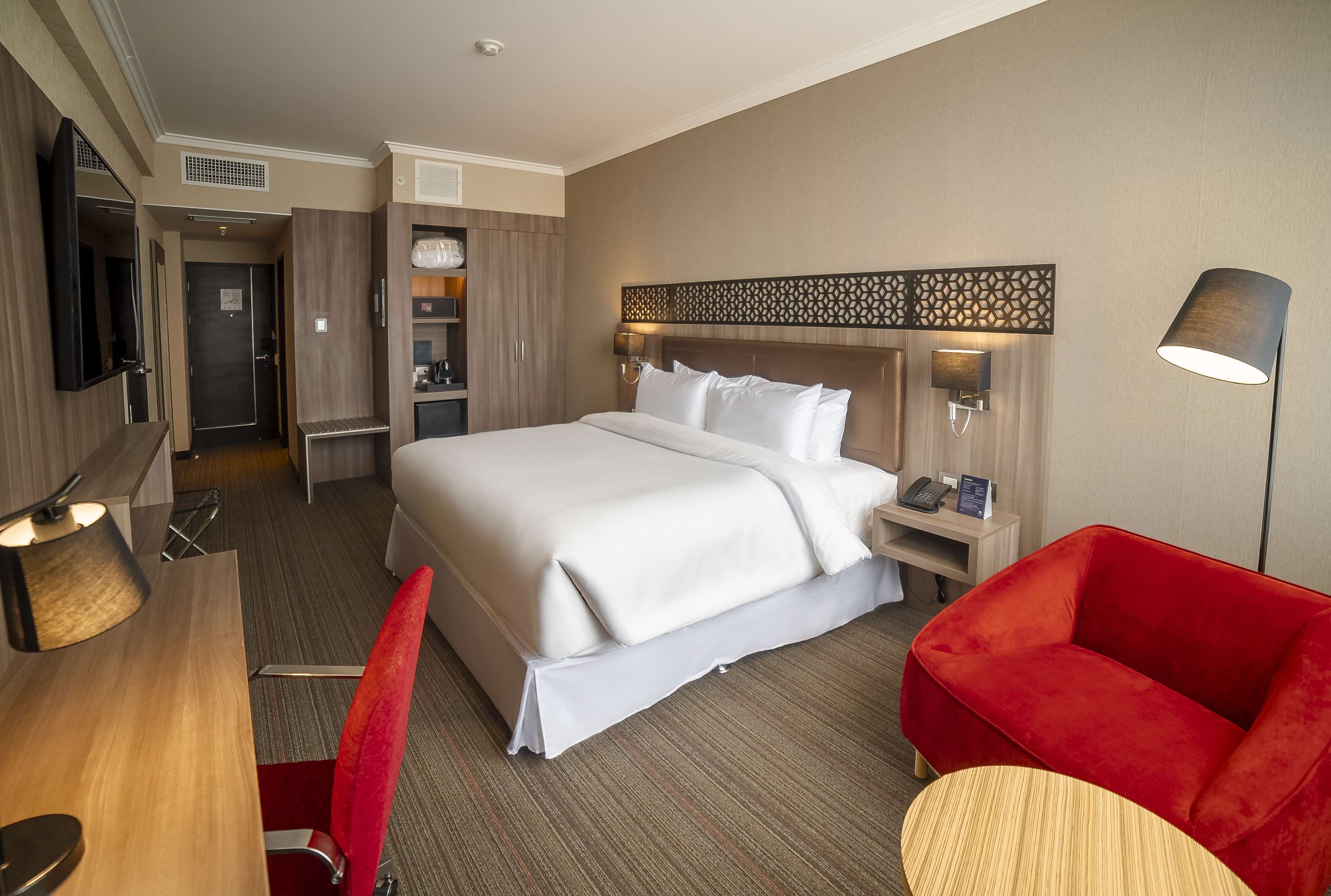 A hotel room with a bed, a desk, a chair, a TV, a lamp, and a red sofa.