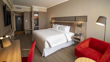 A hotel room with a bed, a desk, a chair, a TV, a lamp, and a red sofa.