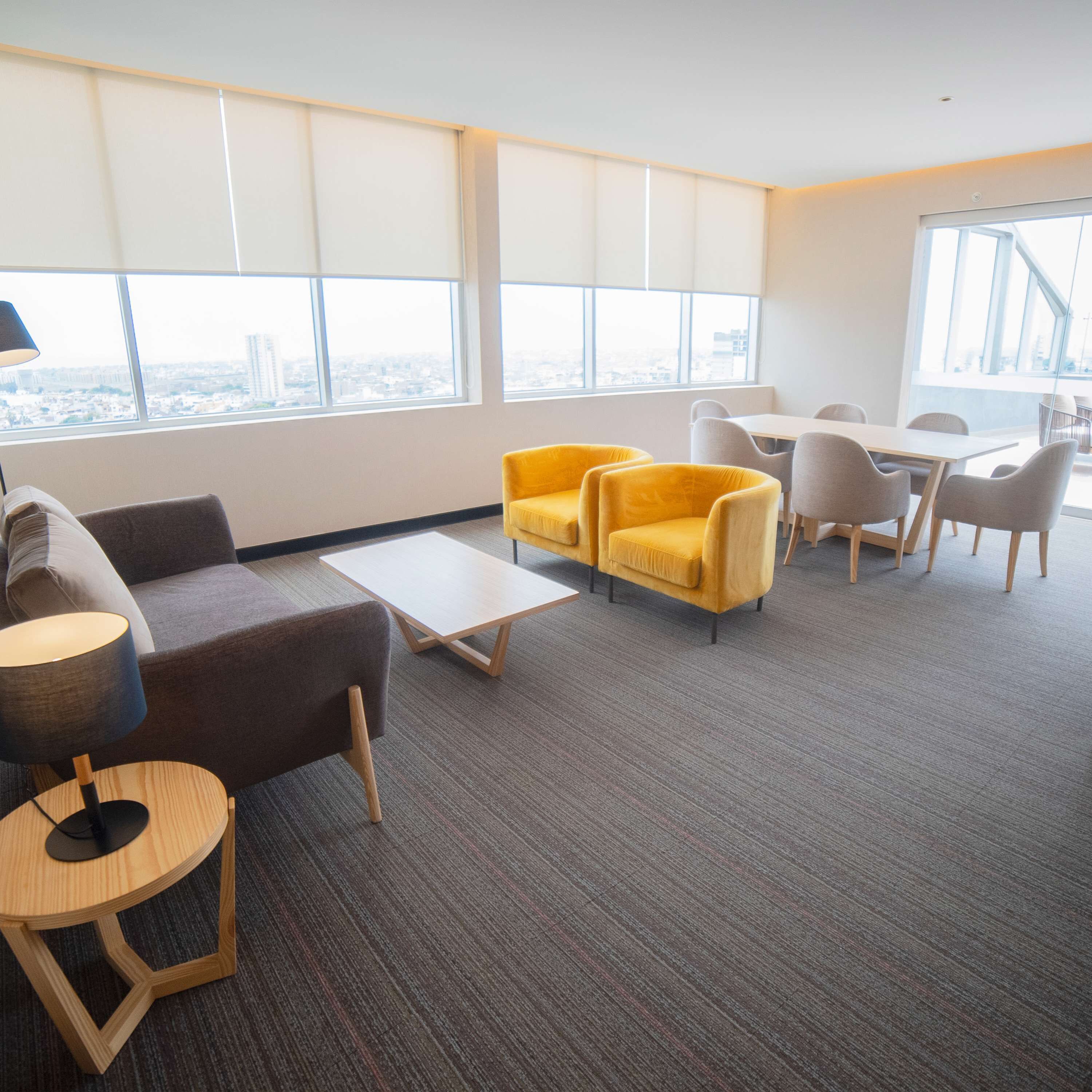 Un espacio de oficina moderno con alfombra gris, un sofá, sillones amarillos, una mesa redonda de madera y una mesa blanca con sillas.