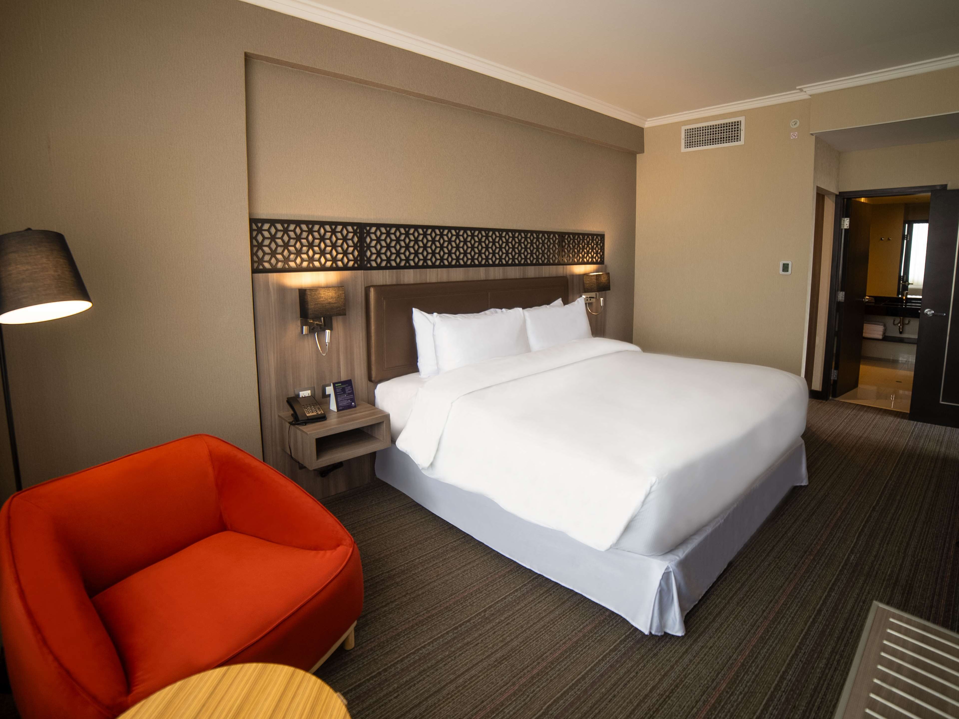 A hotel room with a large bed, a nightstand with a lamp and phone, a red chair, and a small table.