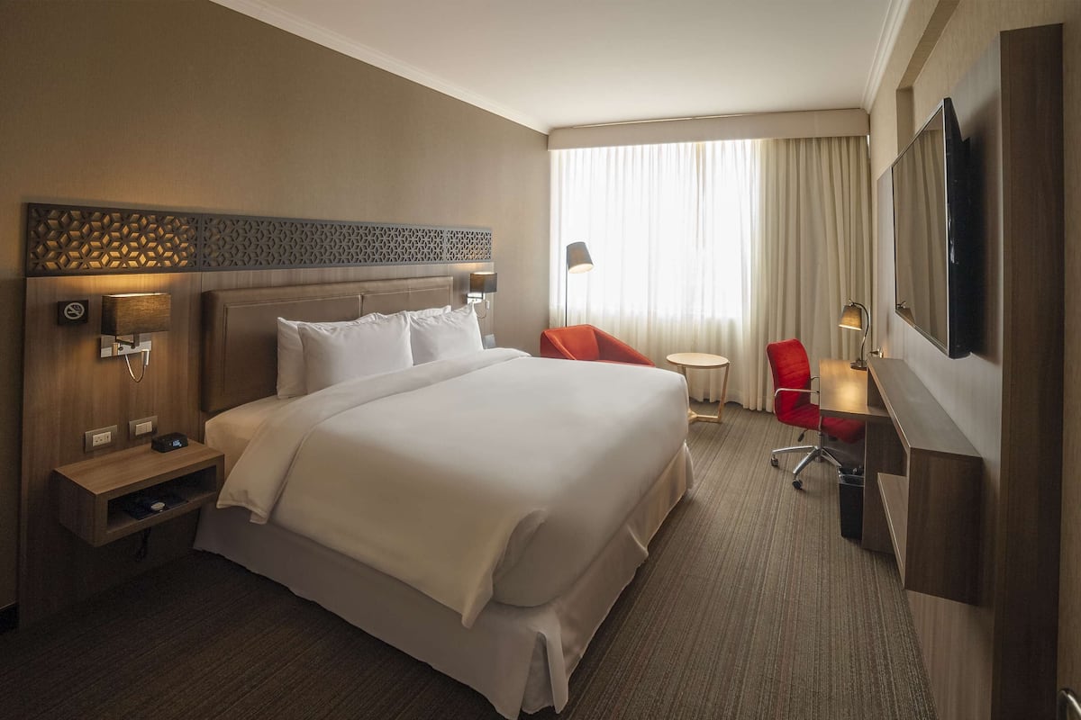 A hotel room with a large bed, a desk, a chair, a TV, and a window with curtains.