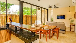 Una cocina moderna con encimera de madera, zona de comedor con mesa y sillas de madera, sofá y un amplio ventanal con vistas a zonas verdes.