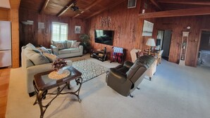 Ein geräumiges Wohnzimmer mit hohen Decken, Holzwänden, einem Sofa, einem Liegesessel, einem Couchtisch und einem Fernseher.