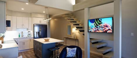 A modern kitchen with white cabinets, a central island, and a built-in refrigerator. There is a dining area with a bar stool and a blue chair. A TV is mounted on the wall above a staircase.
